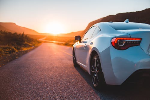 Modern white automobile riding on empty roadway among green plants at sundown time with sun setting on horizon in summer time