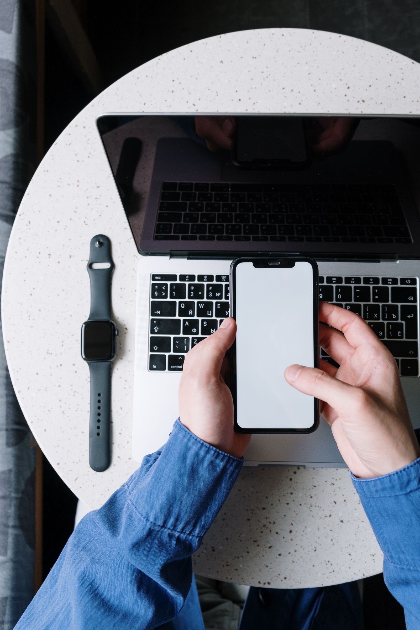 Top view of a person using a smartphone above a laptop, with a smartwatch on the table