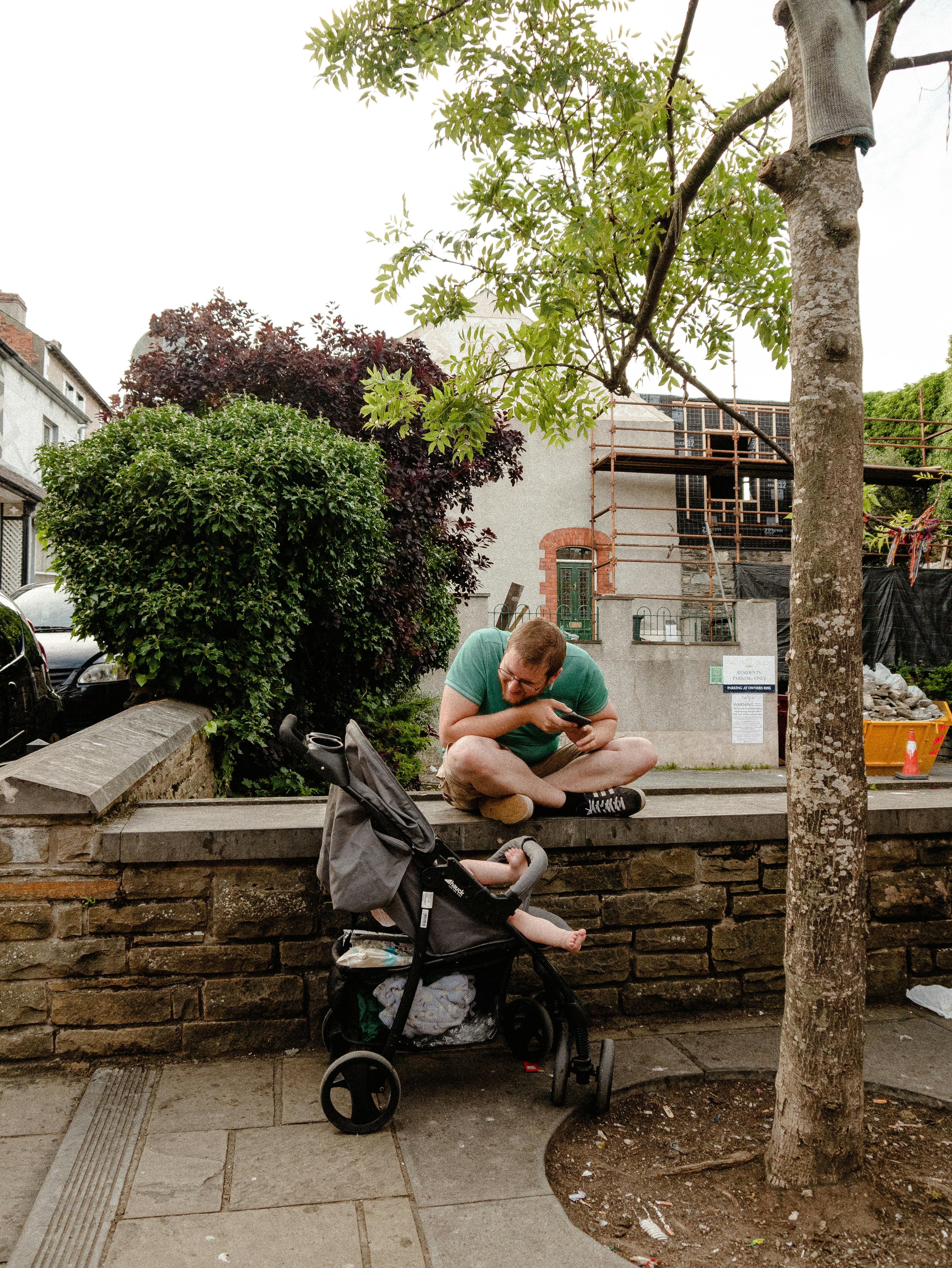 Father sits on a stone wall interacting with child in stroller, urban park scene.