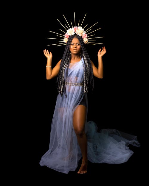 Full body of ethnic female in translucent dress with floral wreath on head standing on black background with arms raised