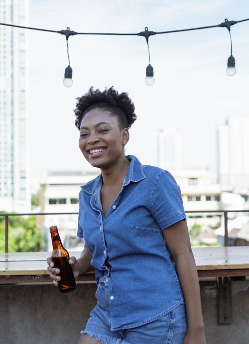 A Woman in Denim Shirt and Shorts Laughing while Holding a Beer Bottle