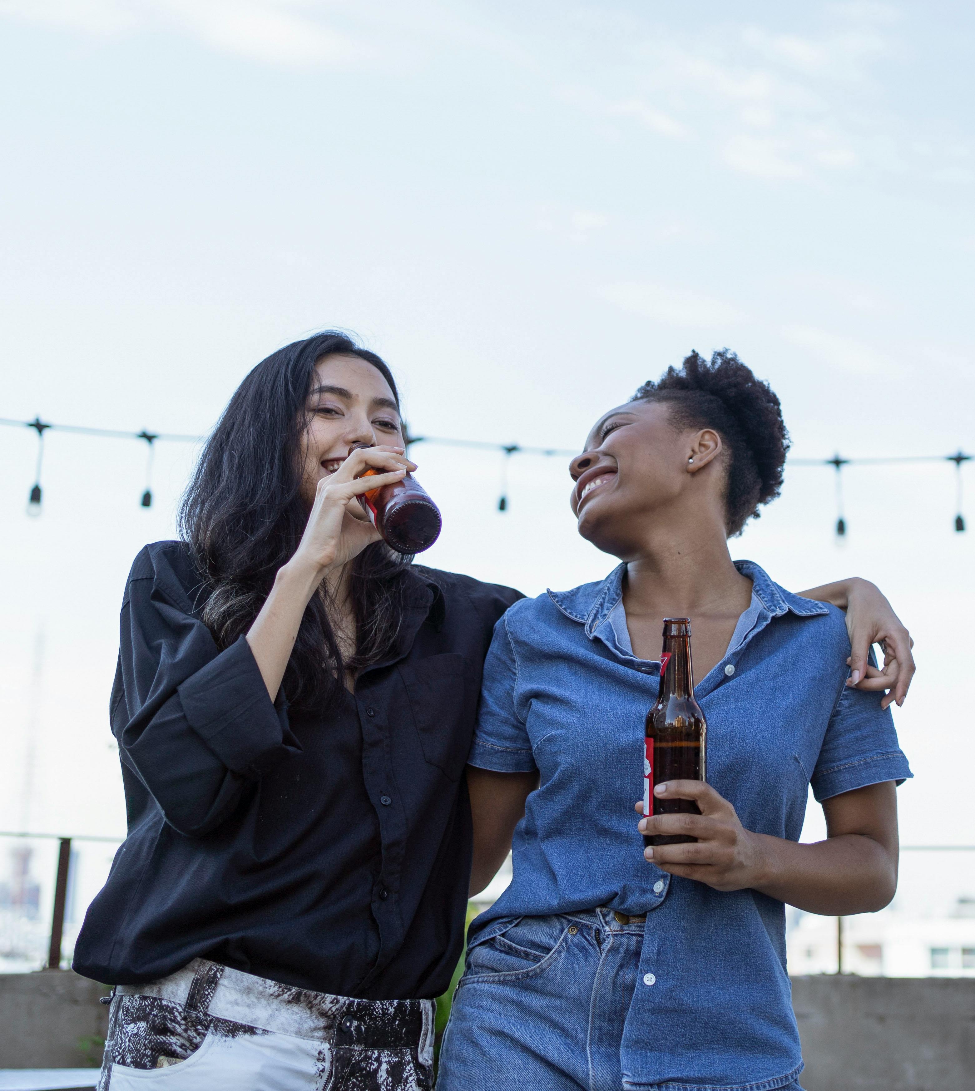 Duas mulheres se abraçam enquanto bebem garrafas de cerveja. Estão rindo e felizes.