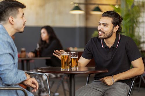 Free Two men enjoying beers and conversation in a relaxed cafe setting. Stock Photo
