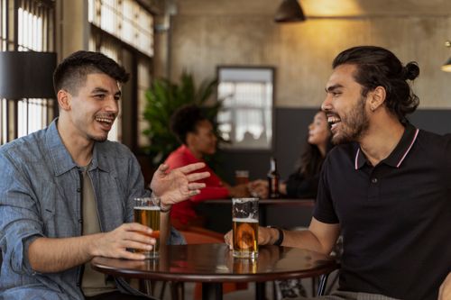 Free Two men laughing and enjoying beers at a café table indoors. Stock Photo