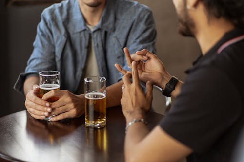 Man in Blue Shirt and Man in Black T-Shirt Talking Over Glasses of Beer