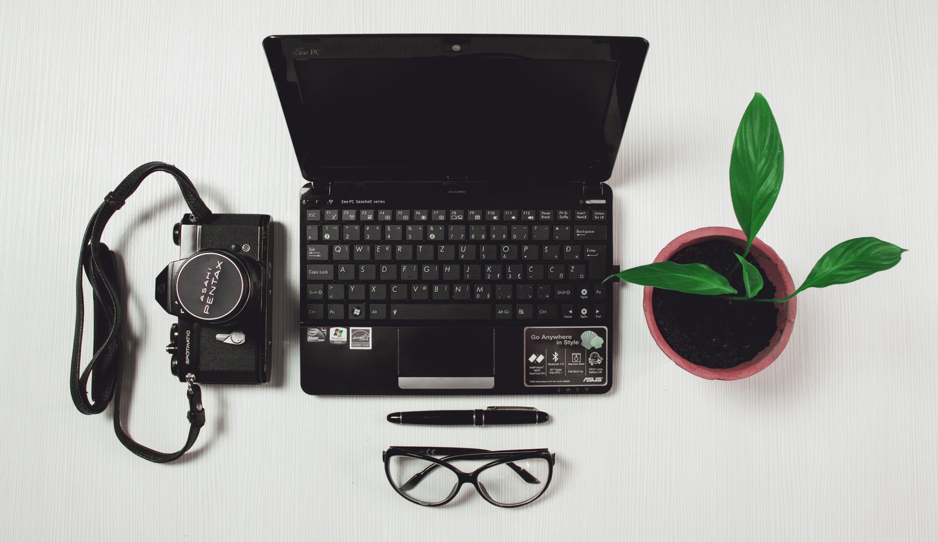 Top view of a workspace with laptop, camera, potted plant, pen, and glasses on a white surface.