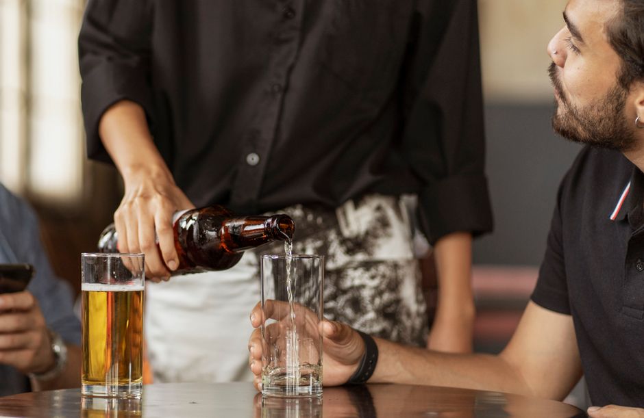 Man with facial hair enjoying conversation as beer is being poured into glasses at a table.