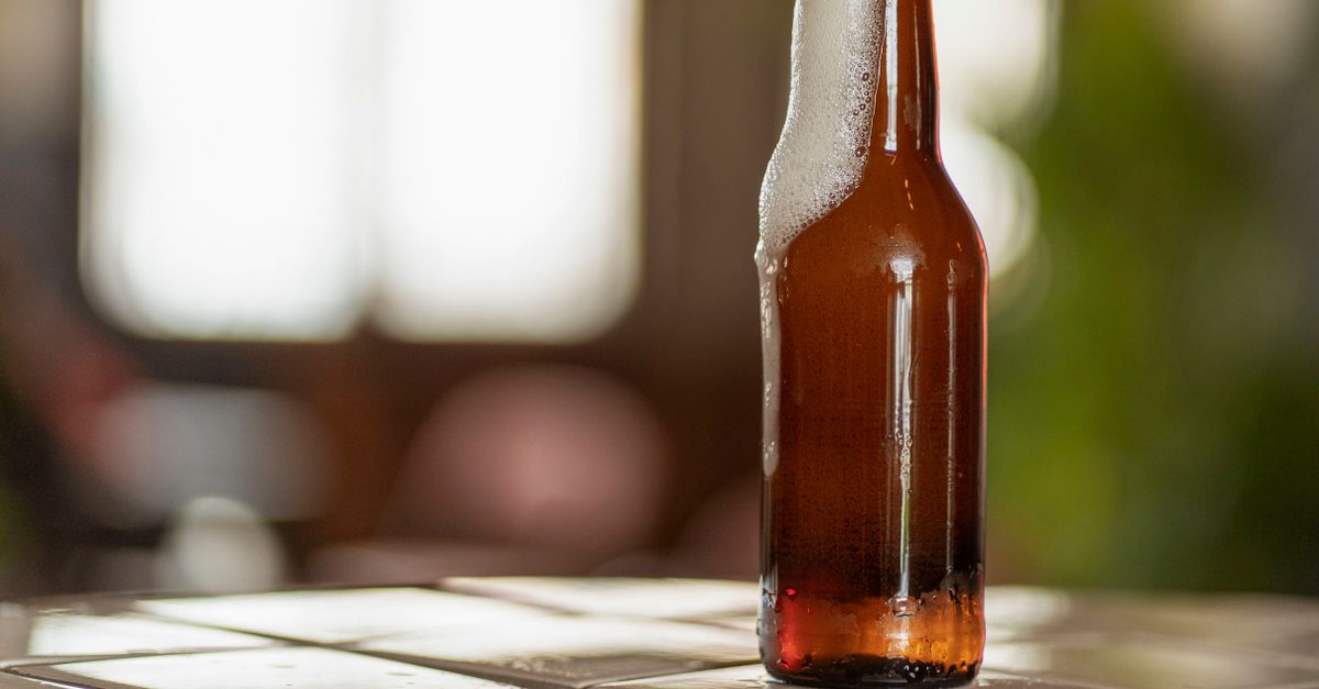 Um close de uma garrafa de cerveja com espuma em uma mesa refletiva, dentro de casa com iluminação suave.