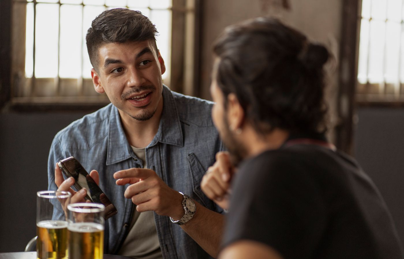 Two men engaged in conversation over drinks at a casual indoor setting, holding mobile phones.