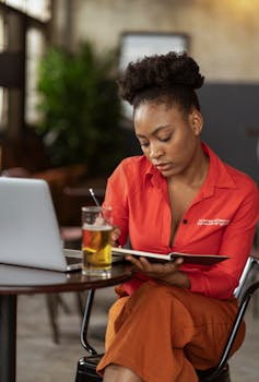 Focused woman in red writing in notebook with laptop and drink nearby in cafe.