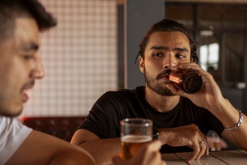 Free Two men having a conversation while enjoying beers at a bar, creating a social and relaxed atmosphere. Stock Photo