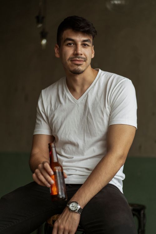 Free Portrait of a casually dressed man holding a beer bottle indoors, smiling gently. Stock Photo