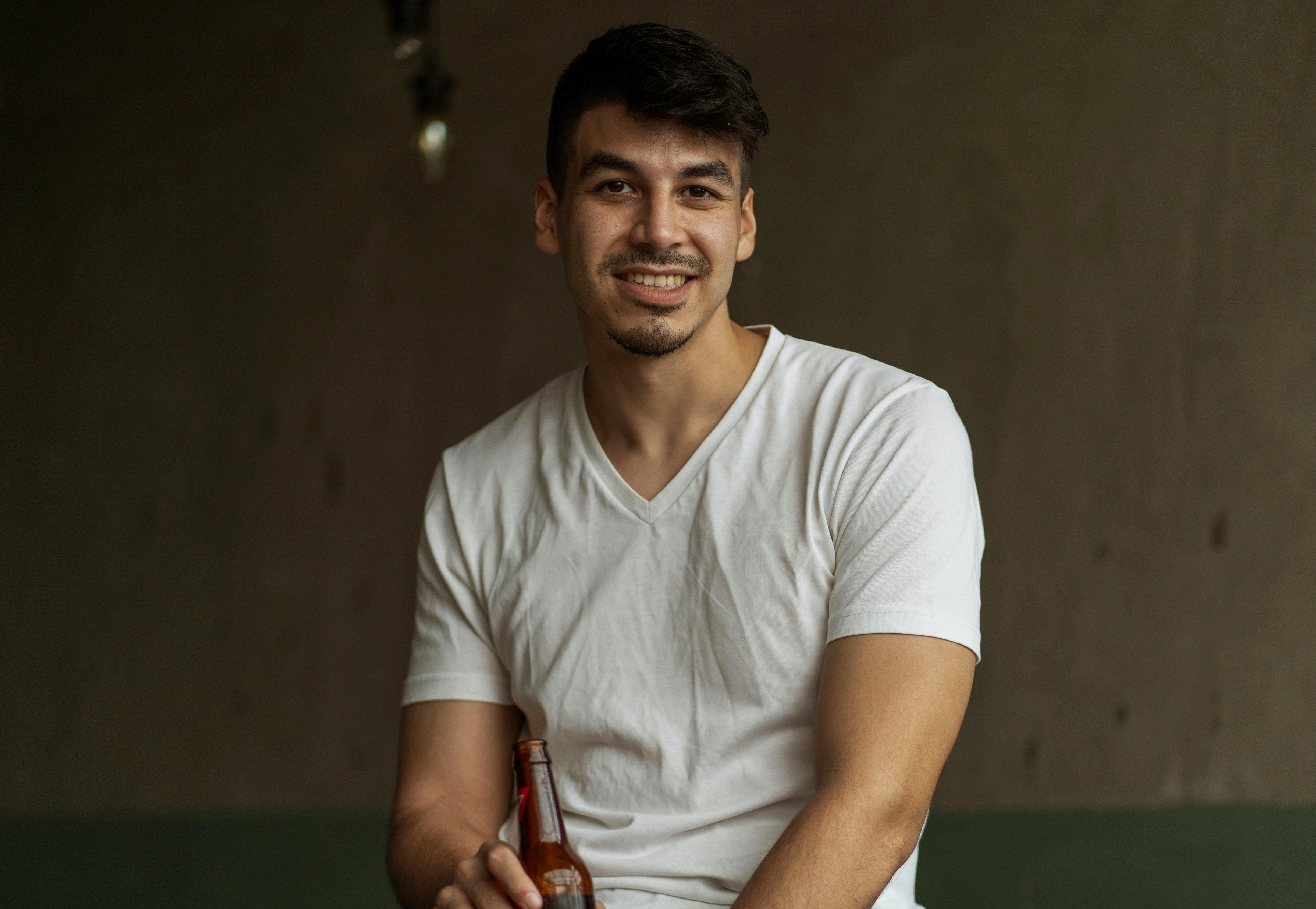 Free Young man with short hair holding a beer bottle indoors, wearing a white t-shirt, smiling. Stock Photo