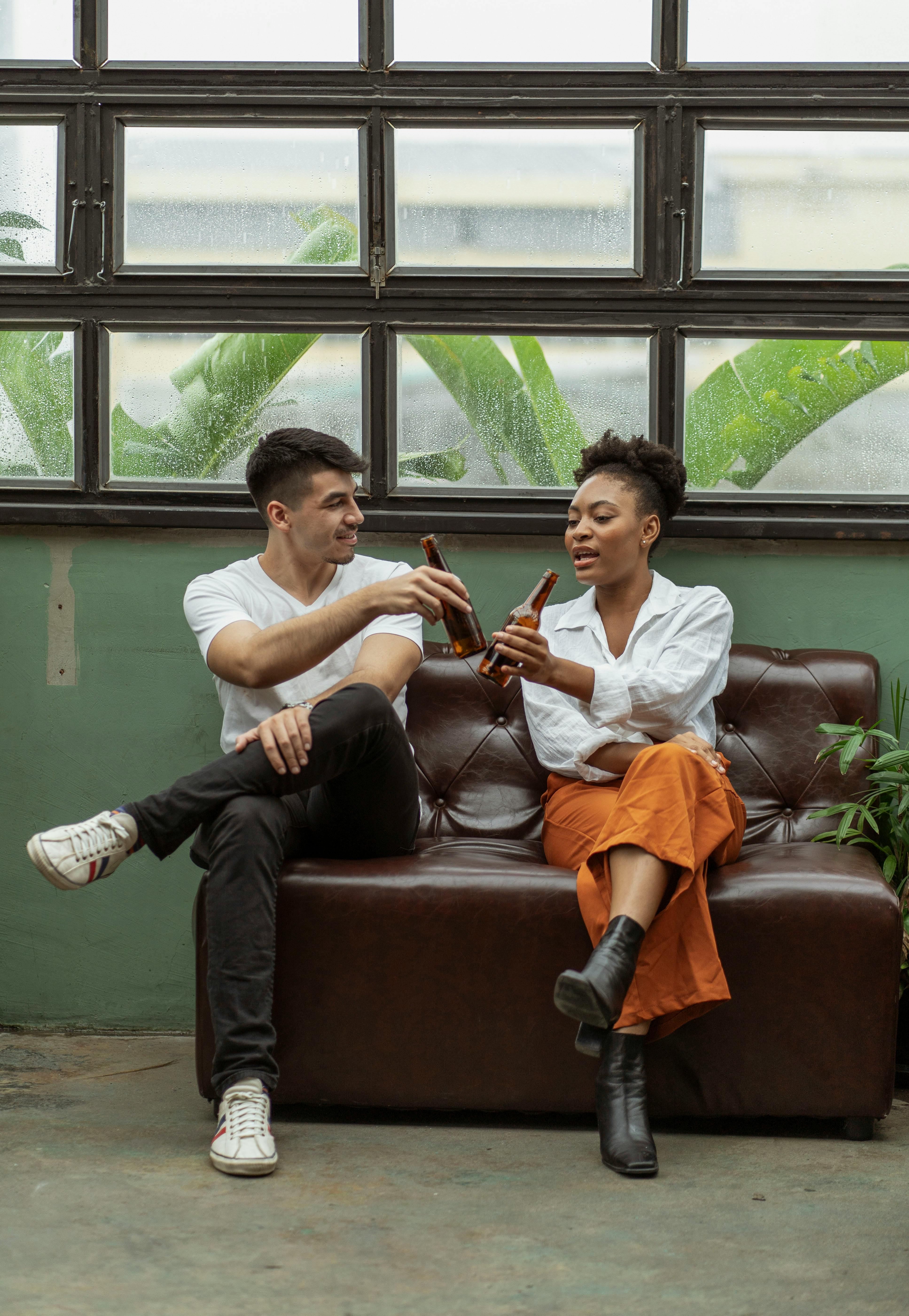 Free Two adults sitting on a couch enjoying drinks in a modern indoor setting. Stock Photo