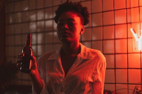 Free A woman in a white shirt holds a beer bottle under moody red lighting in an indoor bar setting. Stock Photo