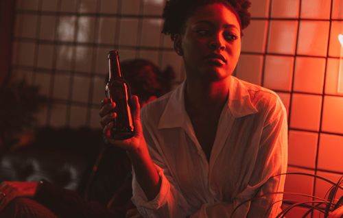 Free A woman sitting indoors holding a bottle under neon red light, creating a cozy bar atmosphere. Stock Photo