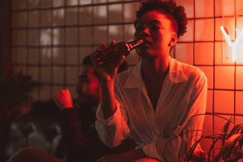 Free A woman in a white shirt drinks from a bottle in a dimly lit club with red neon lighting. Stock Photo