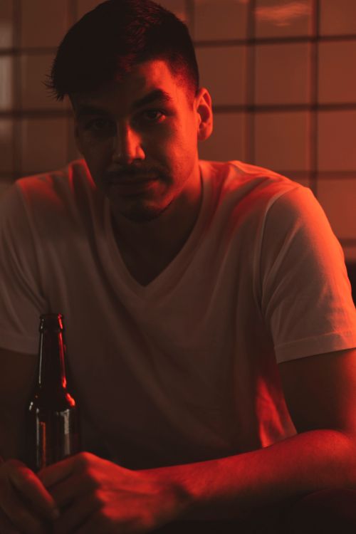 Free A contemplative man with short hair sits in a dimly lit room, holding a beer bottle under red light. Stock Photo