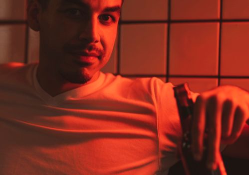 A man in a t-shirt sits relaxed while holding a beer bottle under red lighting.
