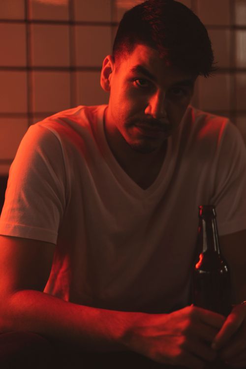Free A contemplative man seated indoors holding a beer bottle under soft red lighting. Stock Photo