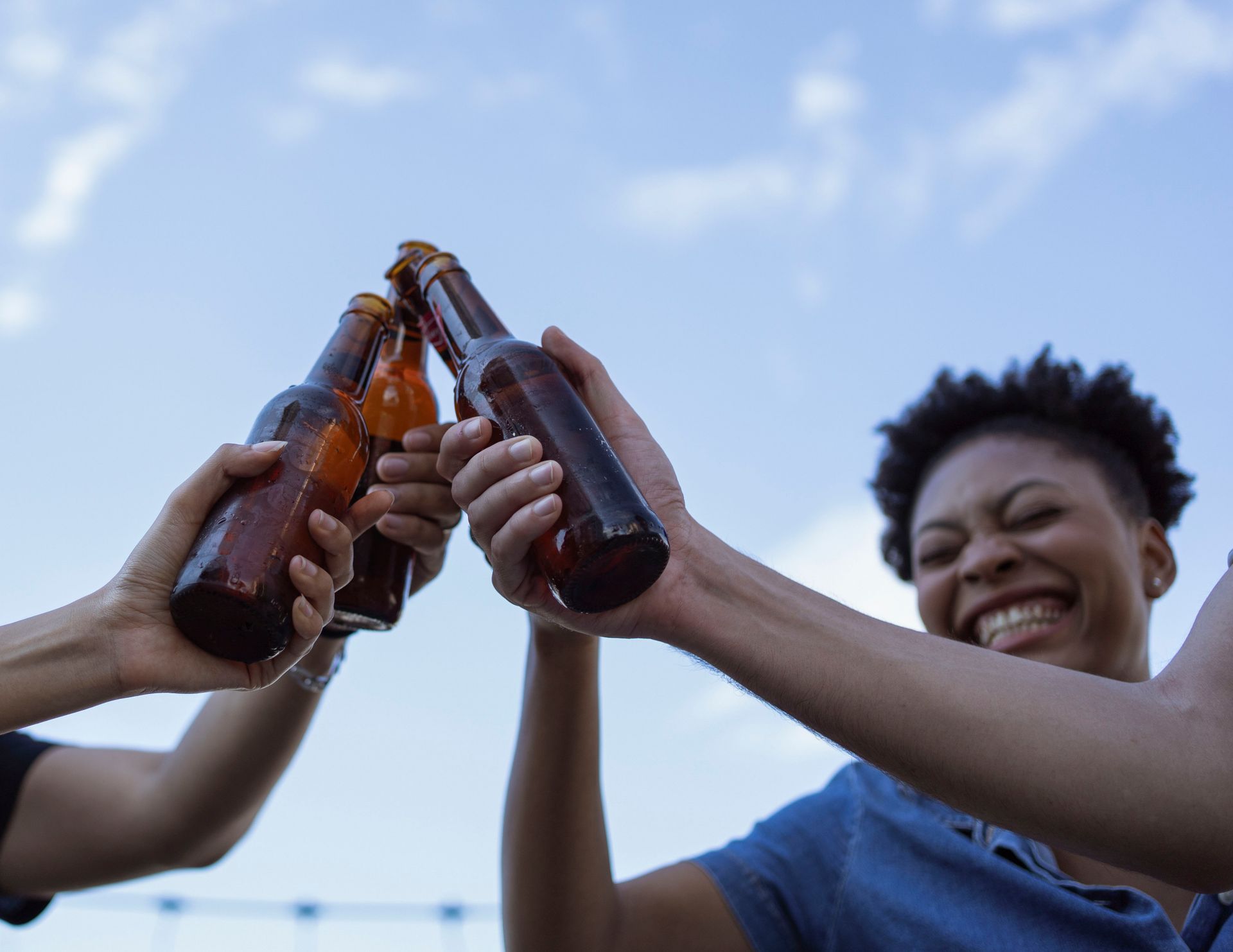 A joyful gathering as friends clink beer bottles under a clear blue sky, celebrating life.
