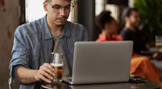 一个戴眼镜的年轻人在咖啡馆里，一手拿着啤酒杯，一手操作着笔记本电脑工作