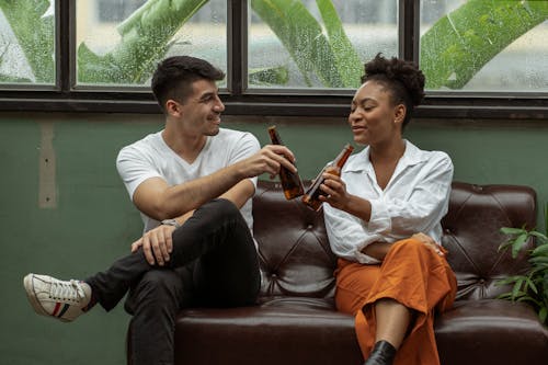 Free Man and Woman Sitting on a Couch and Clinking Beer Bottles Stock Photo
