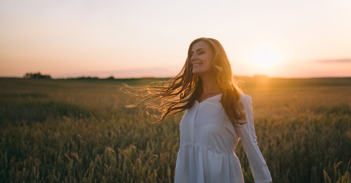 A woman in a white dress smiles joyfully amidst a sunset-lit wheat field.