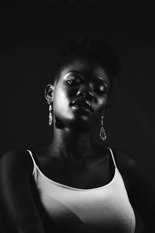 Free Black and white of African American female in white t shirt with earrings looking at camera while standing on black background in studio Stock Photo