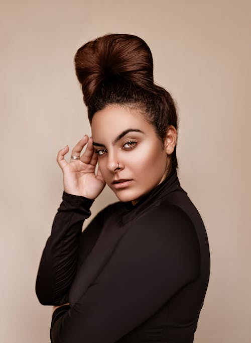 Side view of brown haired female with hairstyle and nose piercing touching face and looking at camera