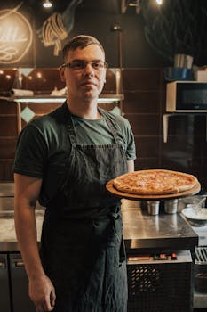 A male chef in a kitchen presenting a freshly baked pizza on a wooden board.