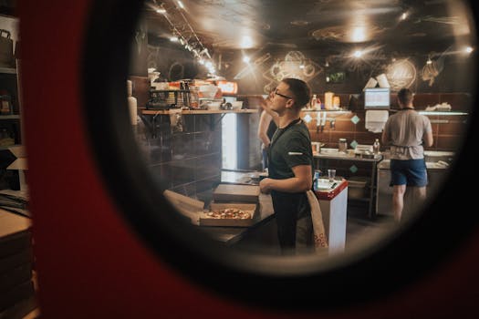 Chefs preparing pizza in a bustling restaurant kitchen through a circular window view.