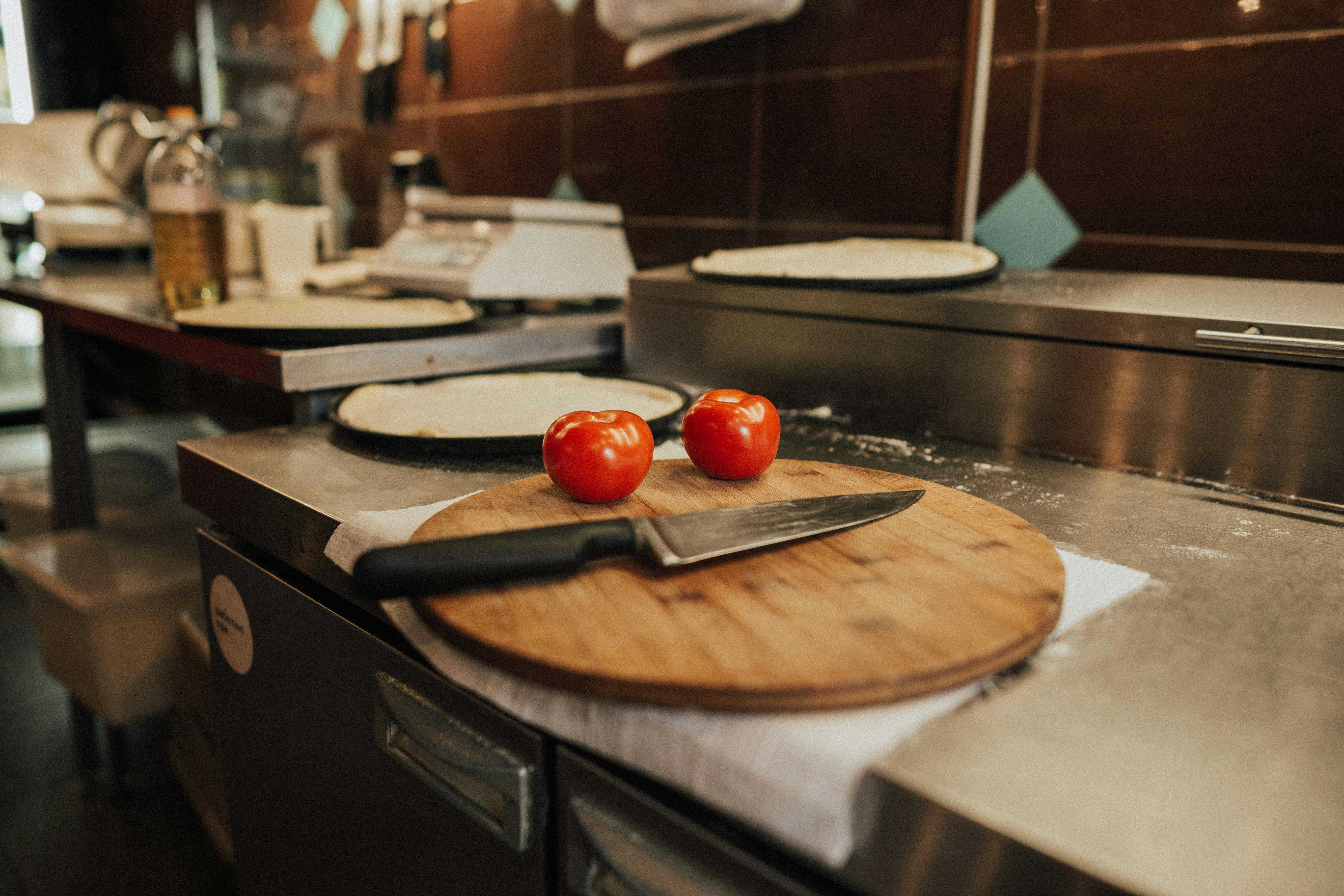 A kitchen scene featuring fresh tomatoes and a knife on a wooden board, set against a counter with pizza dough.