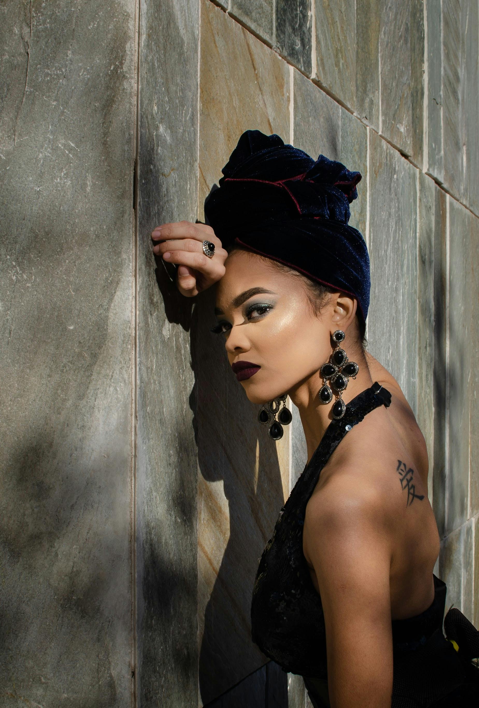 Side view of young sensual tattooed ethnic female in elegant dress and stylish turban leaning head on wall on street and looking at camera