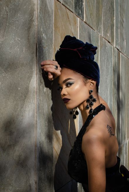 Side view of young sensual tattooed ethnic female in elegant dress and stylish turban leaning head on wall on street and looking at camera