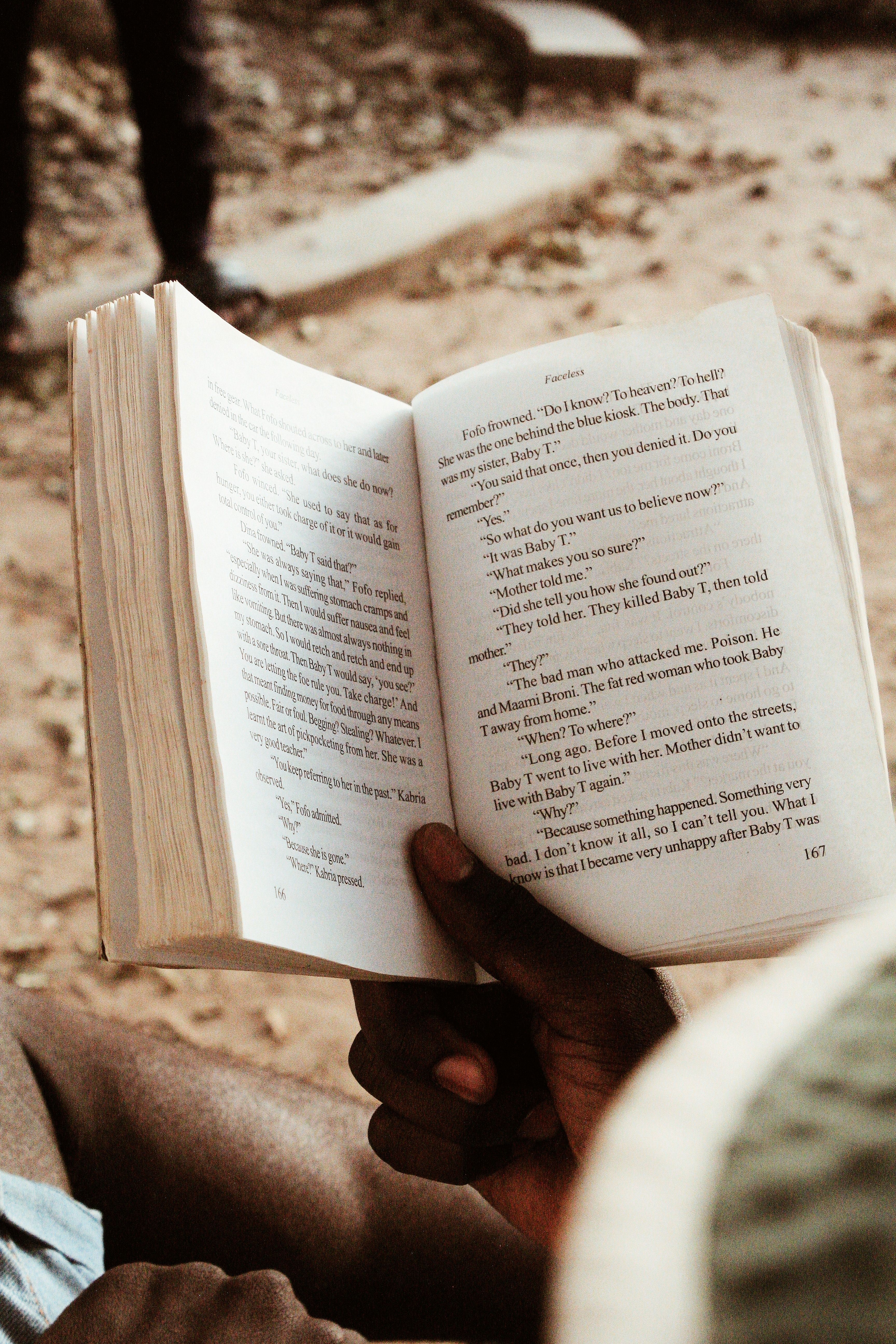 From above of unrecognizable African American male sitting with opened book near stone road while reading in daylight on street in sunny weather