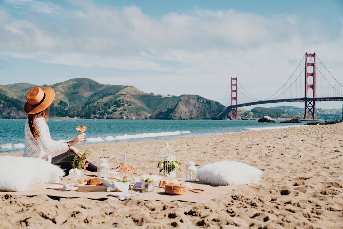 Side view of unrecognizable female traveler in trendy straw hat sitting on sandy beach and drinking wine during picnic near waving sea surrounded by mountains