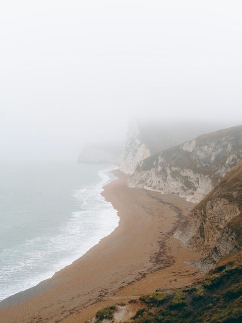 A misty and foggy view of cliffs towering above Durdle Door Beach, England, creating a serene coastal scene.