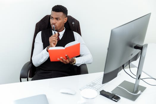 An adult man in a business suit holding a notebook while thinking at his desk.