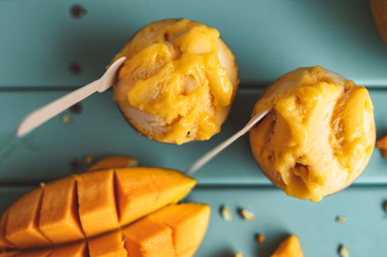 Two scoops of vivid yellow mango sorbet with wooden spoons beside cut mango on teal wood