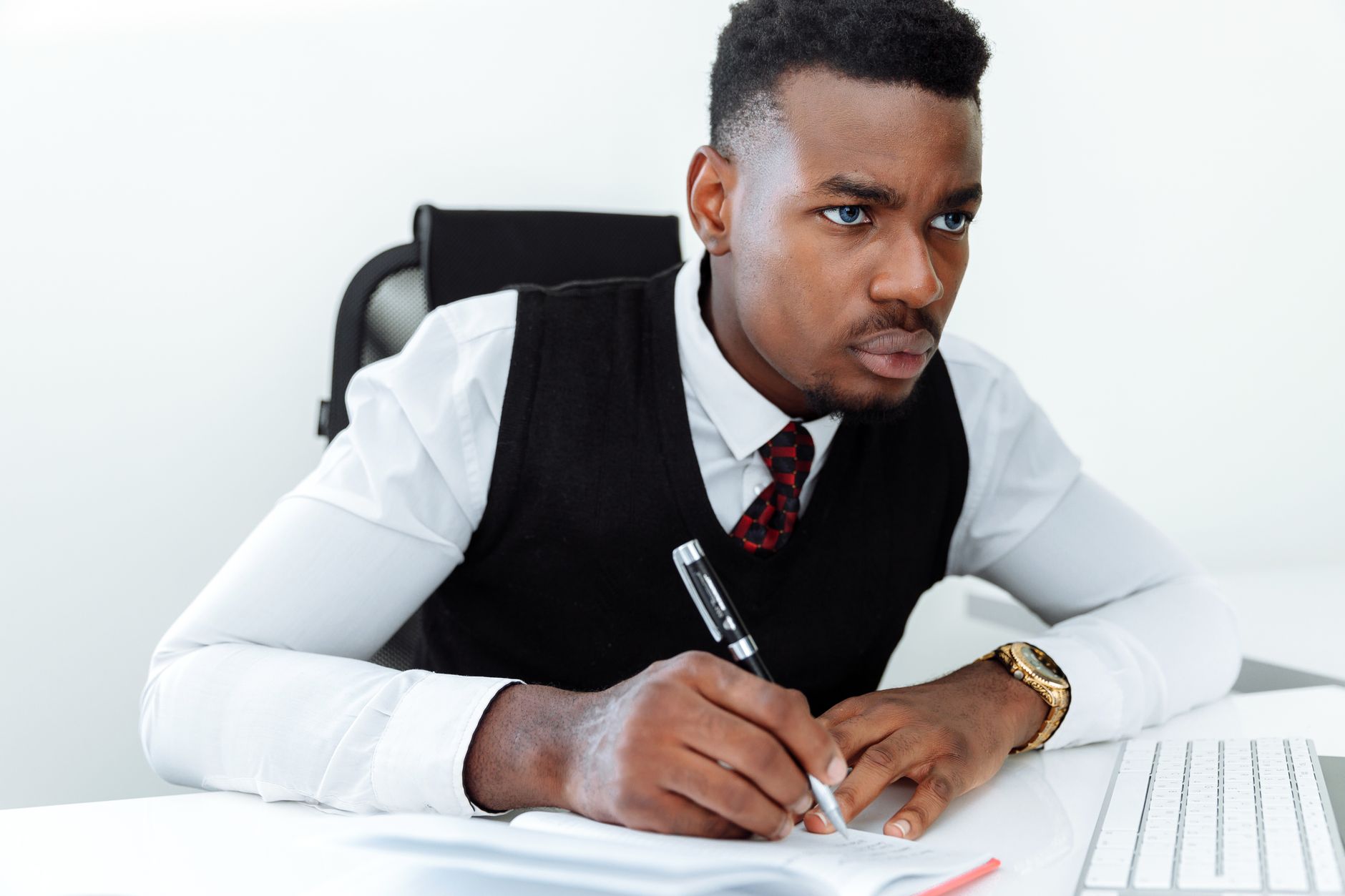 A black adult man in business attire writing at a white desk, focused and attentive.