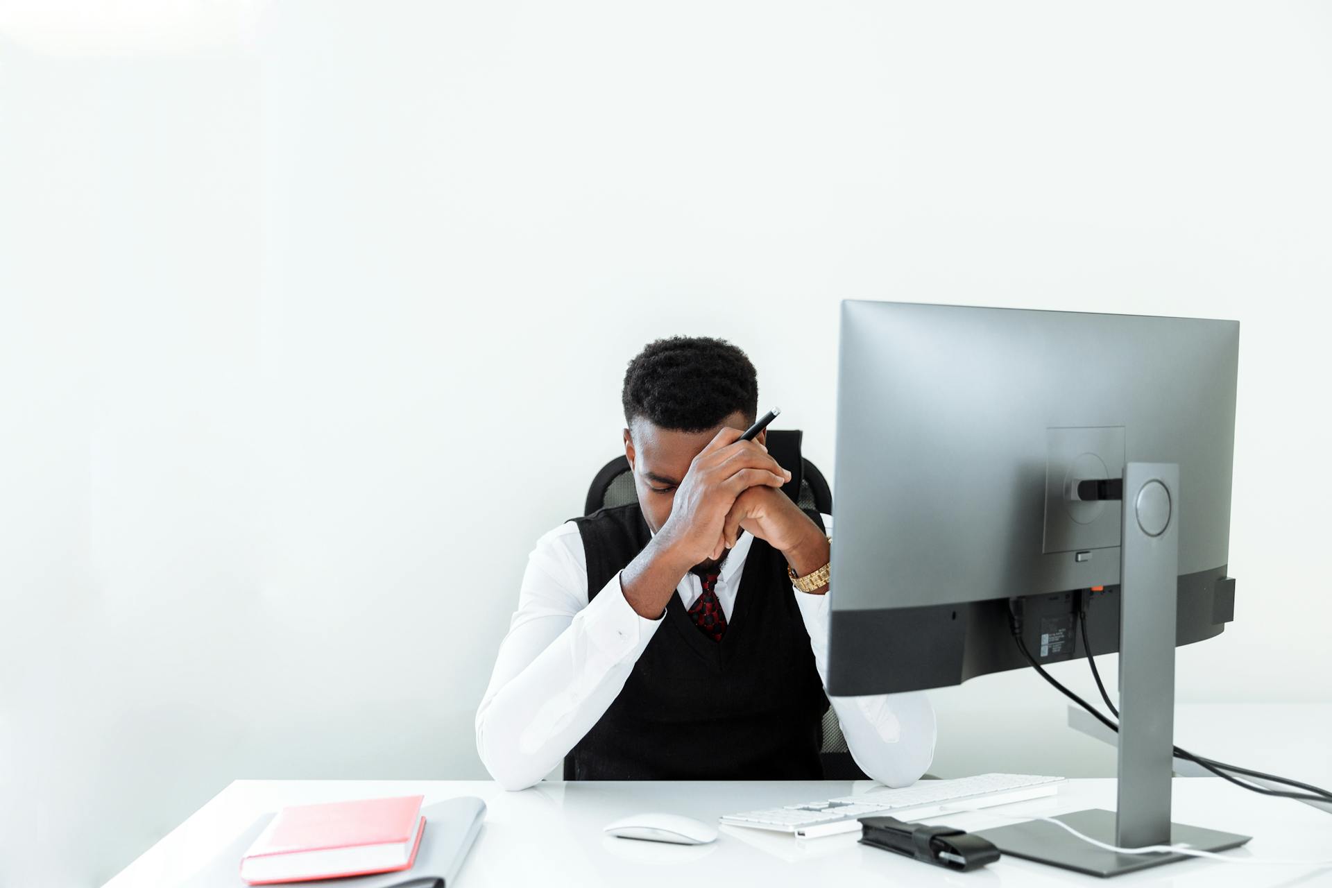 A businessman sitting at a desk, appearing stressed while working at a computer in a bright office.