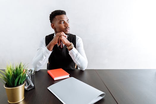 A businessman in formal attire sitting at a desk, deep in thought with office plants and notebooks.