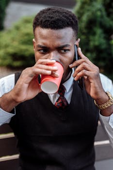 African American man drinks coffee while talking on the phone outdoors.