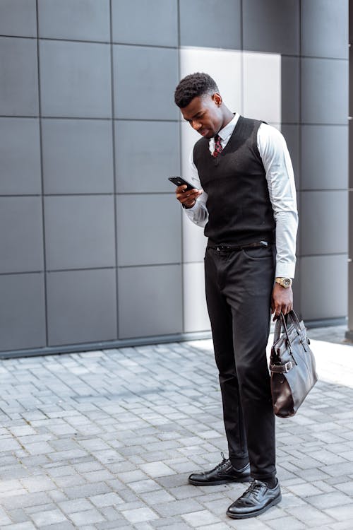 Man in White Long Sleeves and Black Vest Using Cellphone while Walking on the Street