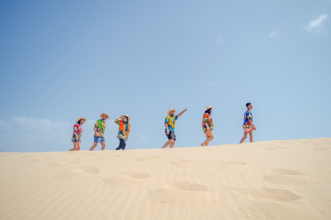 Low angle side view of unrecognizable people in colorful summer clothes walking on sandy beach against cloudless blue sky on sunny day
