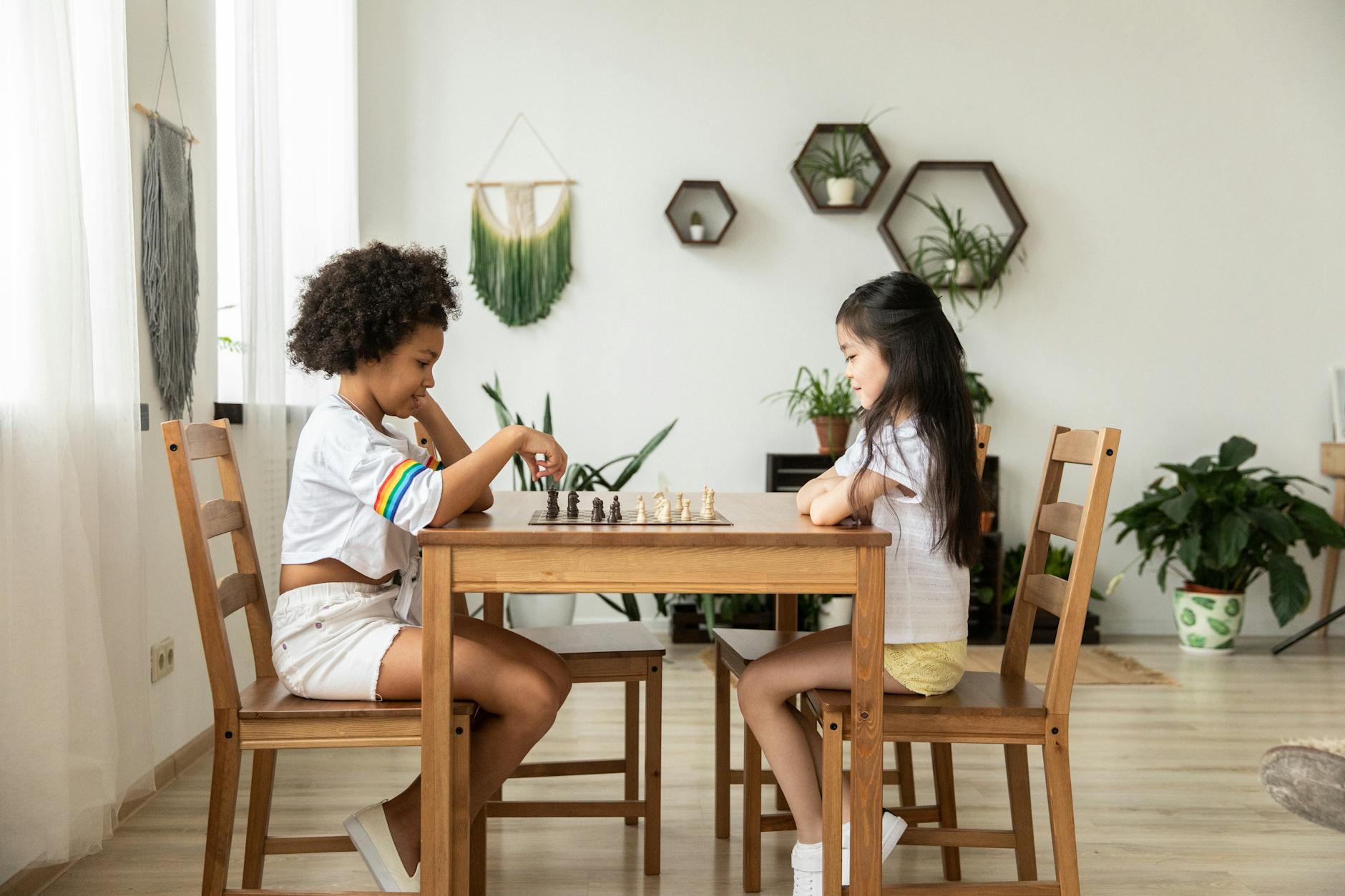 Full body side view of multiethnic girls in casual clothing playing chess at wooden table in bright room decorated with plants in pots