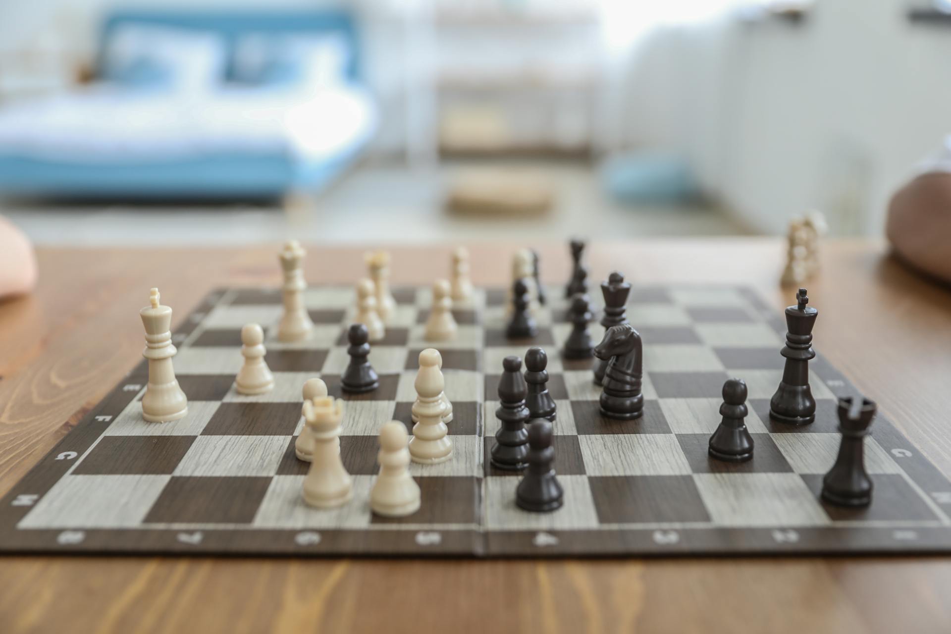 Small white and black figurines on thin square chess board in bedroom in daytime on blurred background