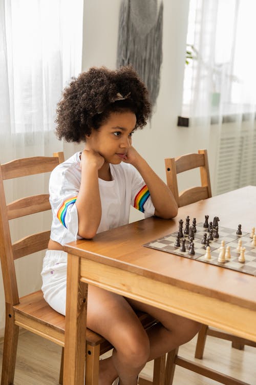 Full body of concentrated African American girl with curly hair touching cheeks while playing chess in daylight