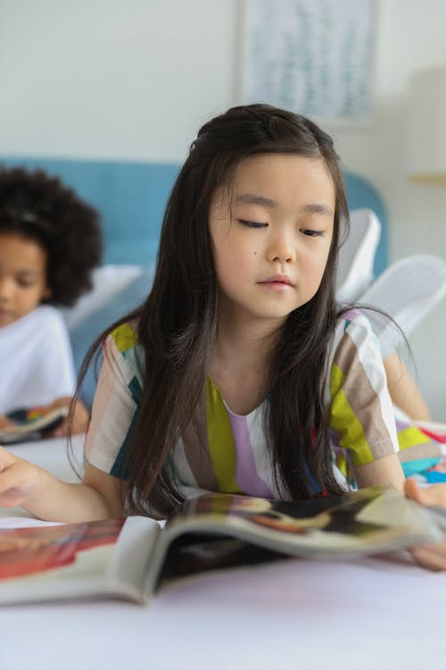 Little cute curious diverse girls lying on bed and watching interesting illustrations in bright journals